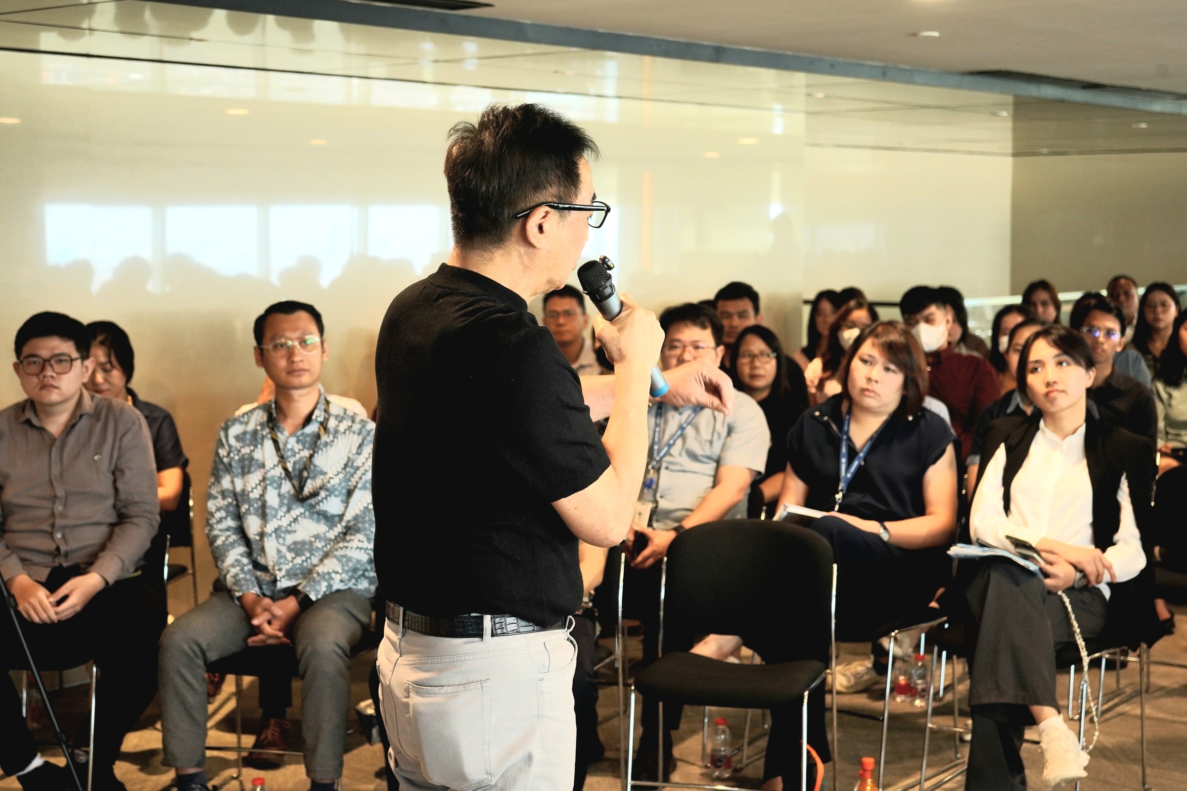 Speaker with a microphone addressing seated attendees in a professional setting.